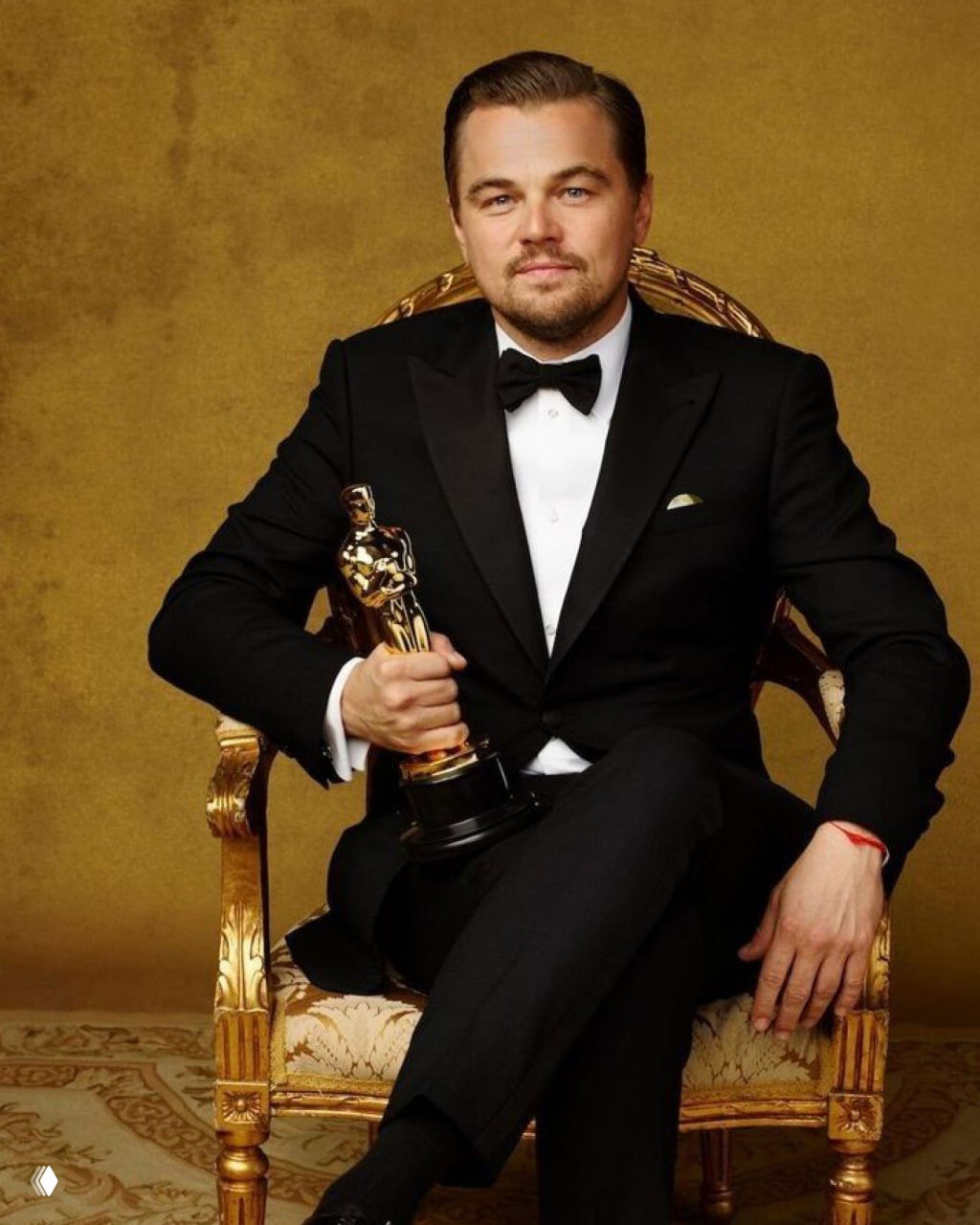 Леонардо ДиКаприо в чёрном смокинге сидит в роскошном кресле и держит статуэтку «Оскар», официальный торжественный кадр со светлым фоном.