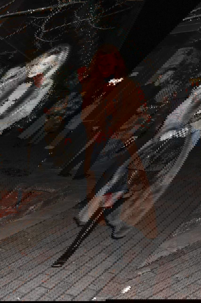 Та же модель в длинной бежевой шубе поверх черного кружевного платья, идет по брусчатке курортного городка в вечернем освещении.