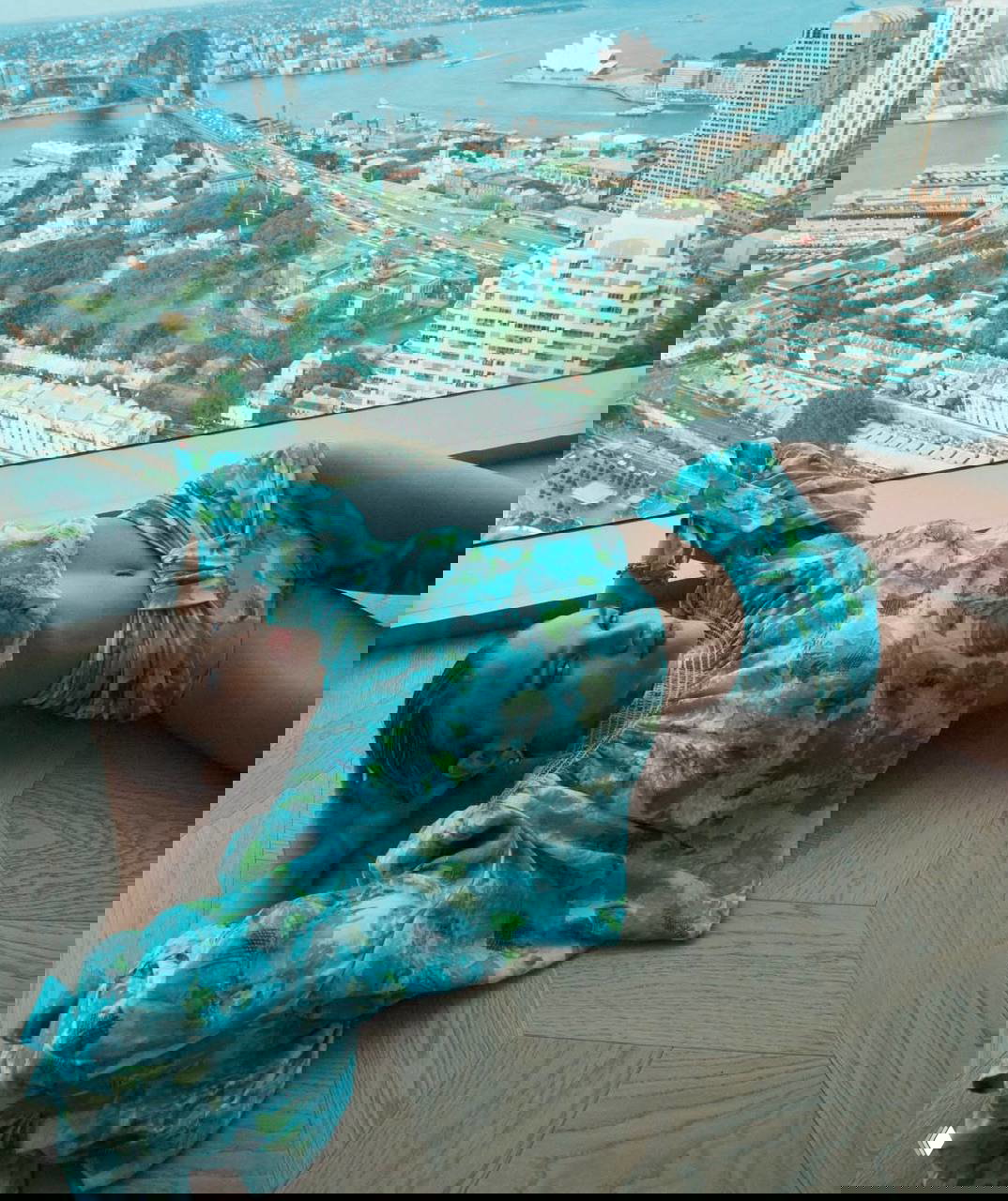 Роузи в голубом цветочном костюме у большого окна с видом на город и Сиднейский оперный театр, расслабленная поза и интерьер квартиры.