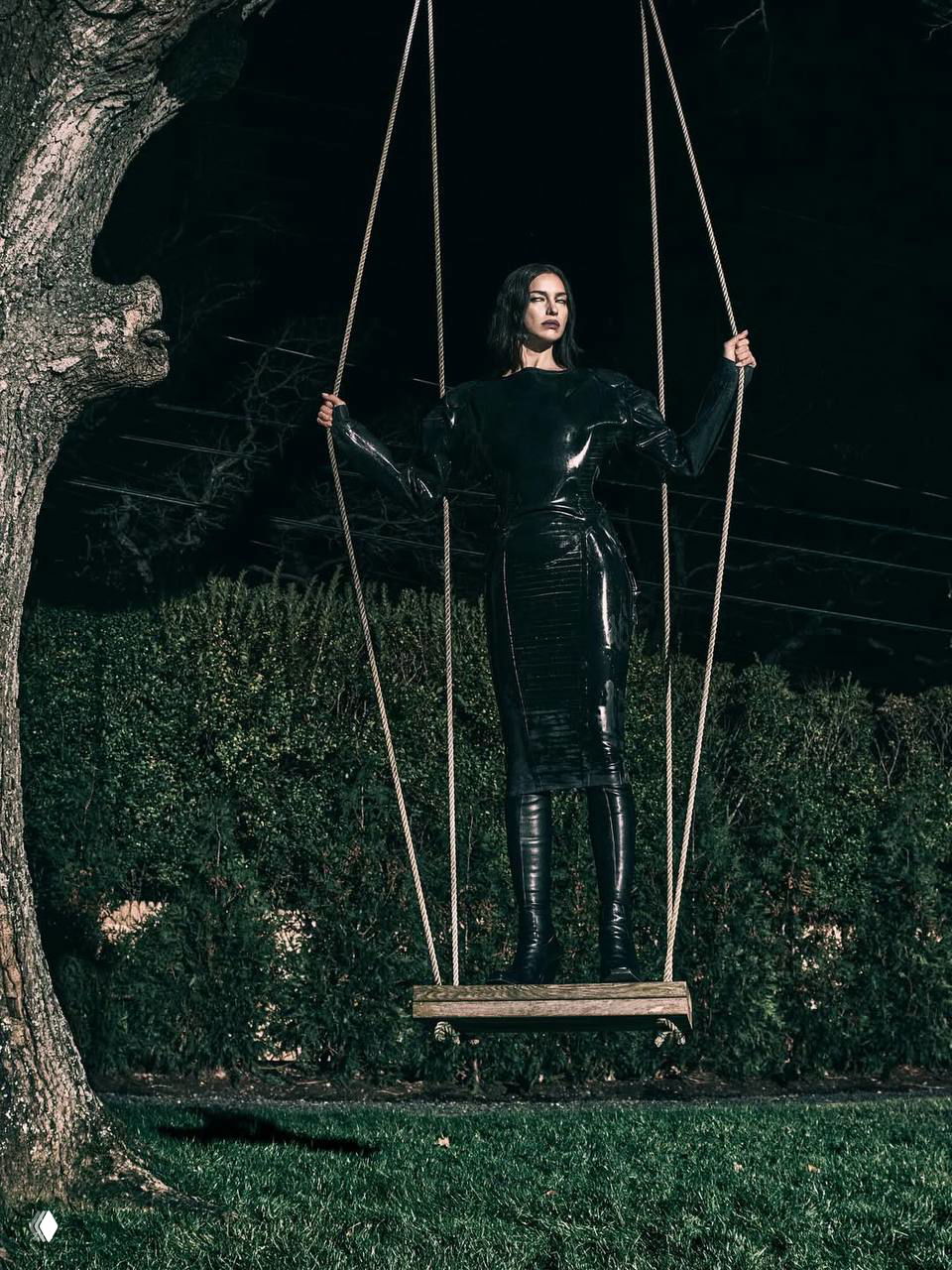 Модель в лаковом платье и высоких сапогах стоит на большой качели в саду ночью — атмосферный снимок с готическими мотивами и контрастной подсветкой.
