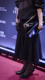 Кадр с красной дорожки: Юлия Снигирь в чёрном миди‑платье держит лакированный клатч, видны туфли на каблуке и баннеры премьеры на фоне.