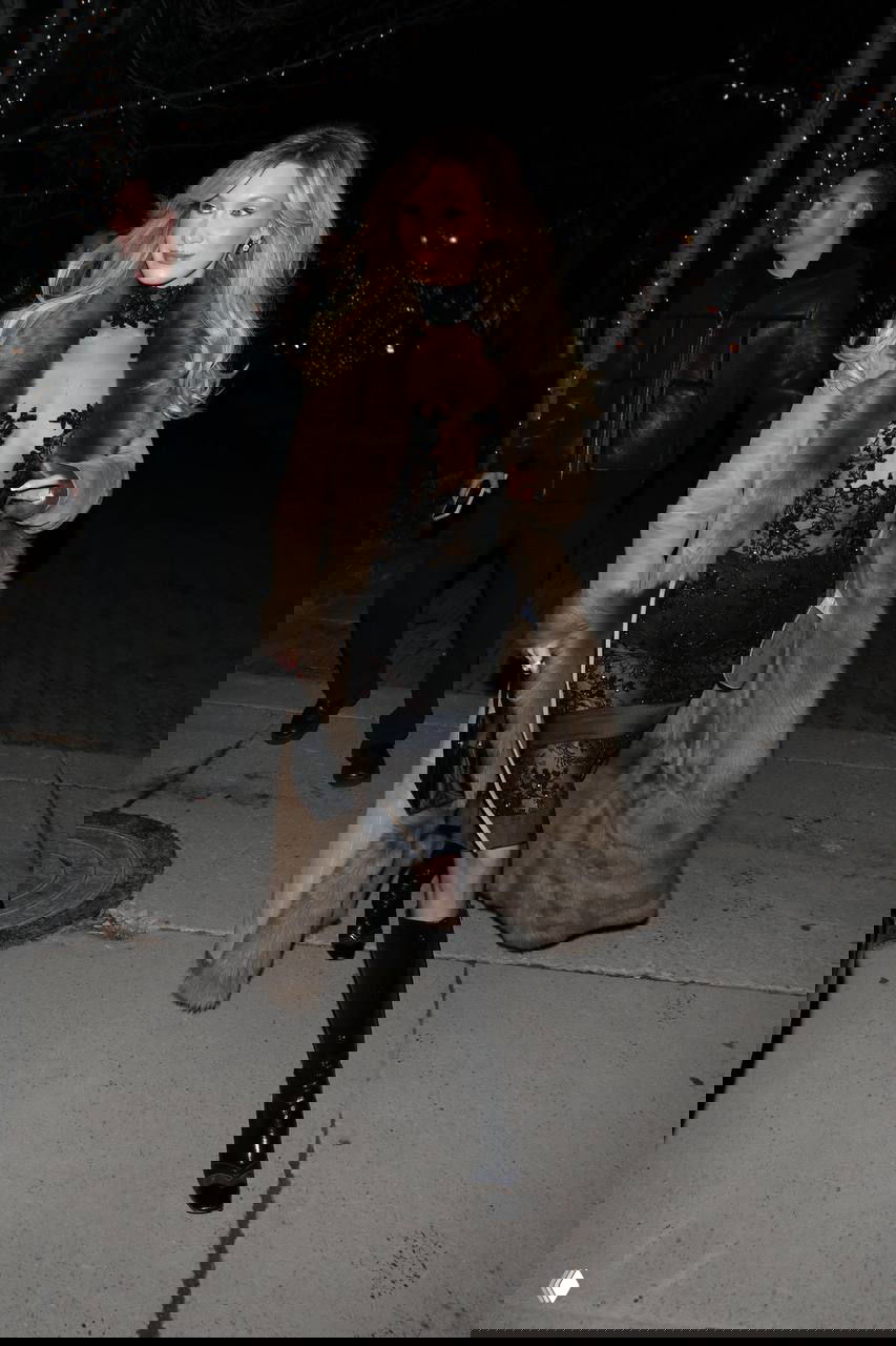 Модель в открытом кружевном платье и высоких сапогах идет по тротуару ночного городка с праздничной иллюминацией на деревьях.