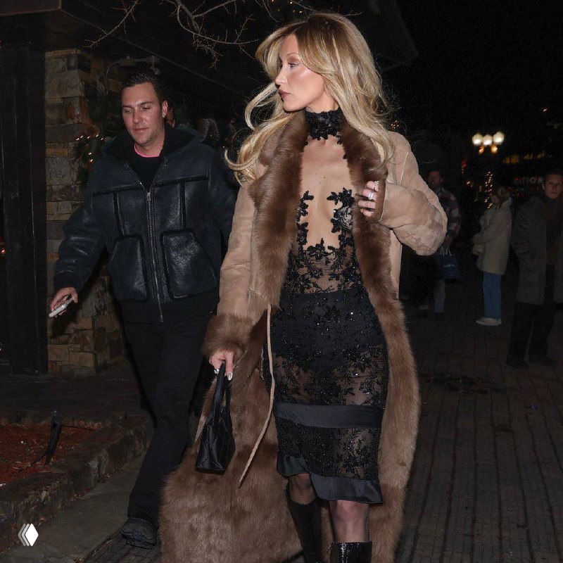 Крупный план модели в бежевой шубе и полупрозрачном кружевном платье держит сумку, на заднем плане прохожие и ночной курорт.