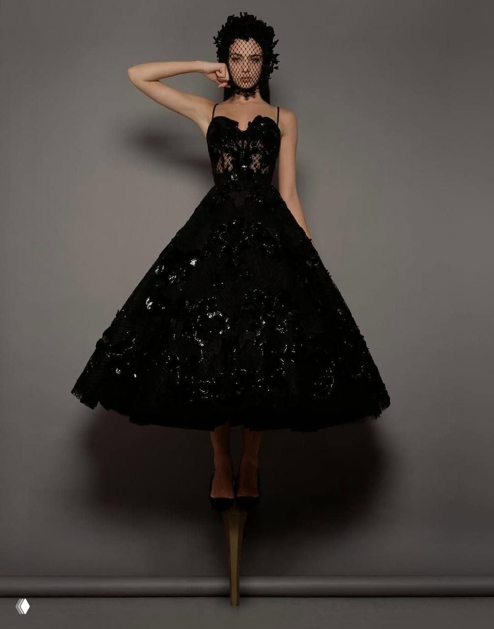 Модель в чёрном кружевном платье с короткой линией подола и высокими платформами, позирует в драматичном фэшн‑стиле на сером фоне студии.