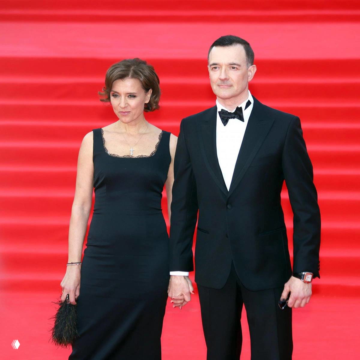 Женщина в черном платье и мужчина в смокинге стоят на красной дорожке у ступенчатого фона, держатся за руки и позируют фотографам.