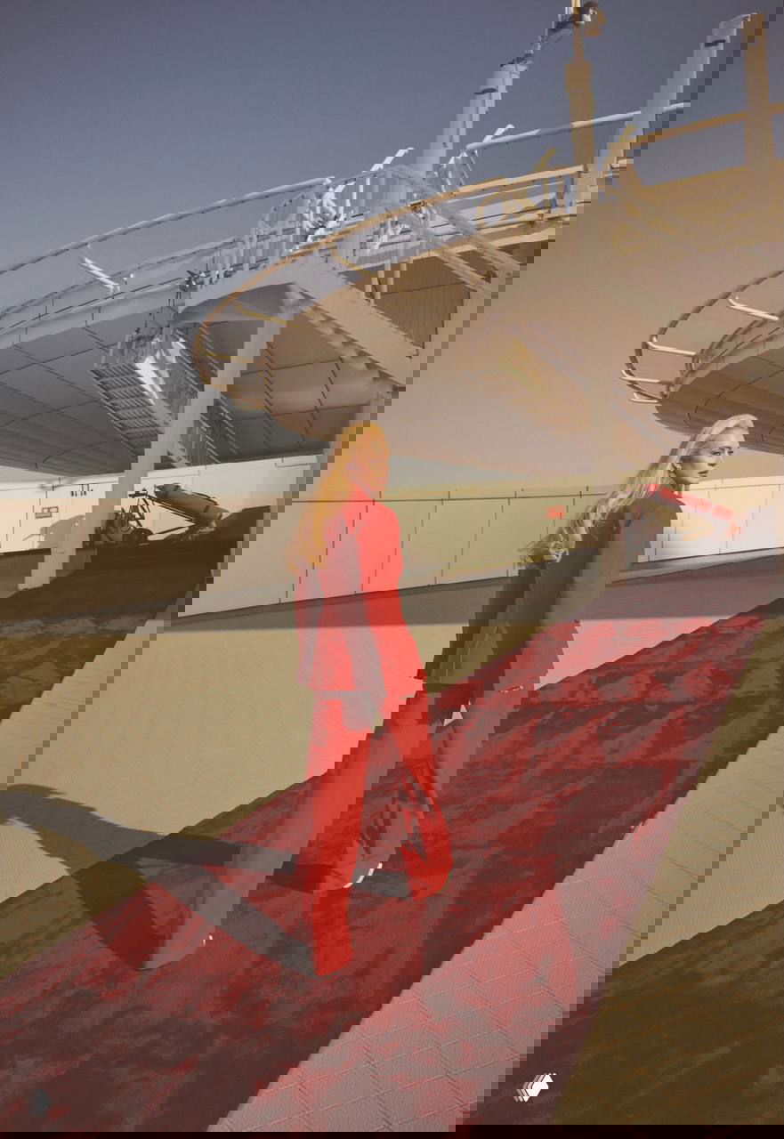 Линдси Лохан в красном костюме идёт по бордовому ковру на крыше с видом на город и архитектурный ангар — динамичный кадр фотосессии.