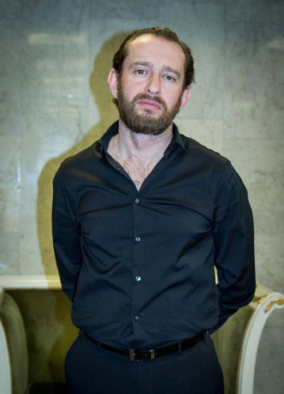 Фотография мужчины в тёмной рубашке на нейтральном фоне, поза стоя, мрачный или серьёзный выражение лица, интерьерный снимок.