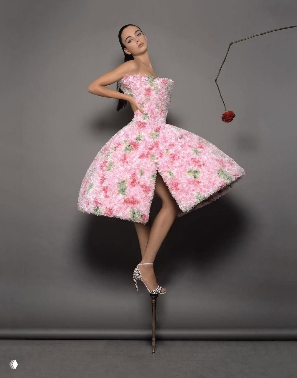 Модель в розовом объёмном платье с текстурой, напоминающей цветочные помпоны; поза на декоративной шпильке в художественной студийной постановке.