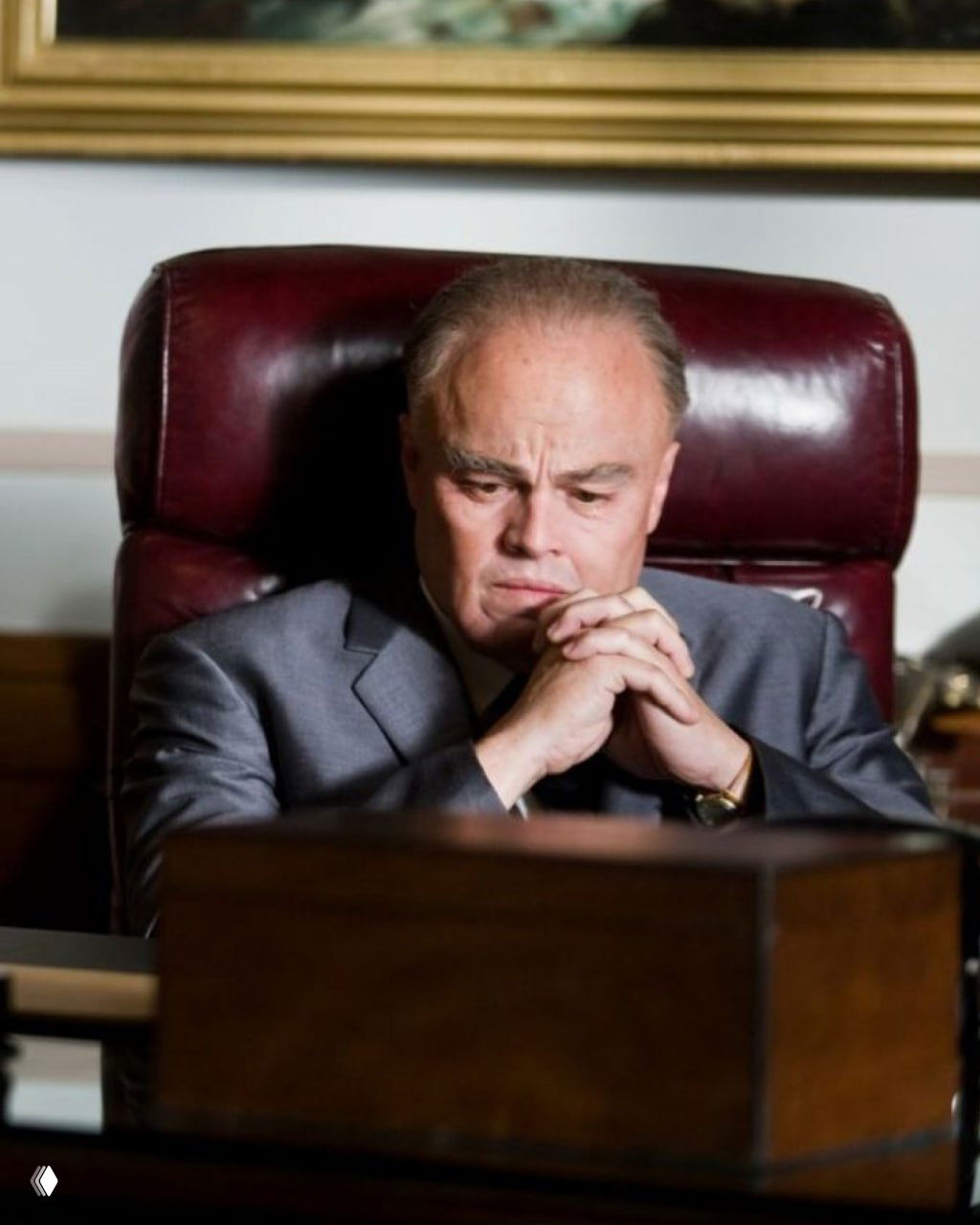 Кинематографический кадр с ДиКаприо в образе зрелого персонажа за письменным столом, строгий костюм и задумчивое выражение лица.