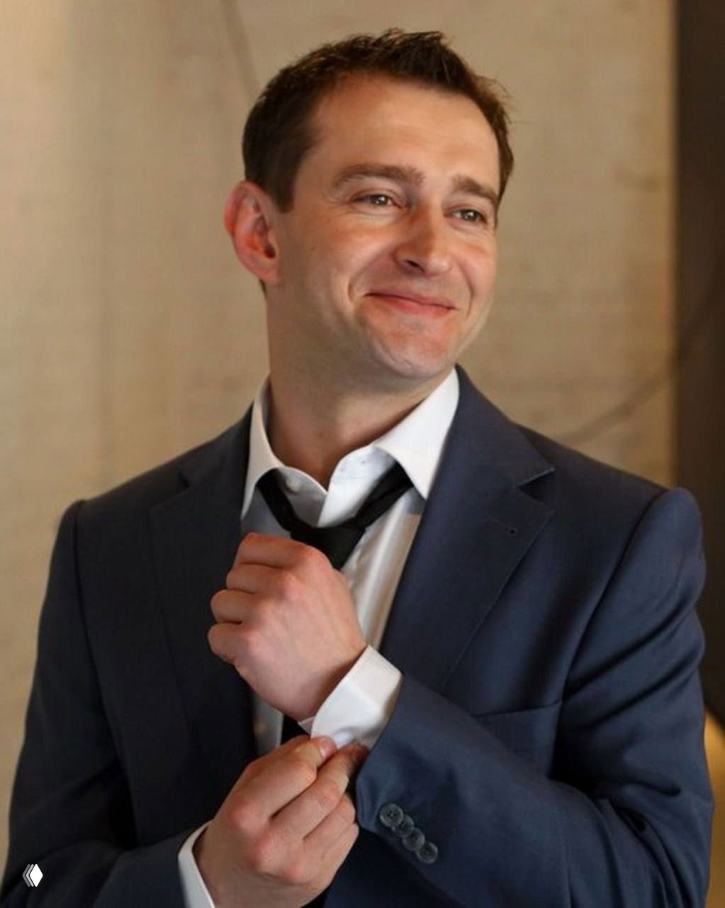 Деловой портрет мужчины в пиджаке и галстуке, улыбающийся образ, полупрофиль, тёплое освещение и нейтральный интерьер.