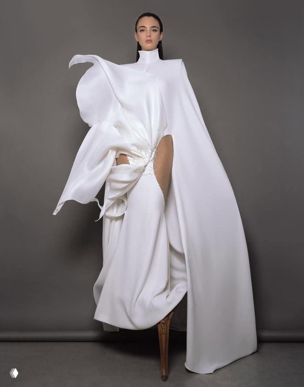 Модель в белом асимметричном платье с драпировками и высоким разрезом, позирует в статичной художественной композиции на сером фоне.