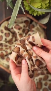 Крупный план кусочка блина с леопардовым рисунком в руках, выделен контраст пятен; на заднем плане видно зелёное растение и домашняя обстановка.