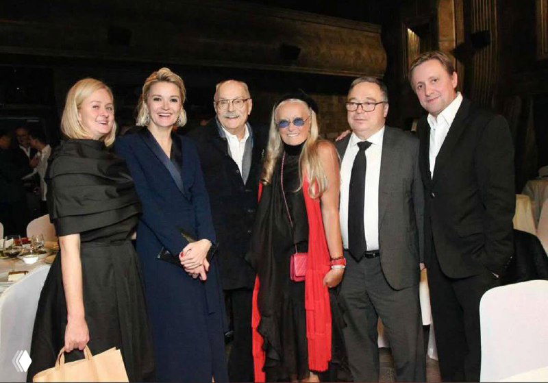 Ряд приглашённых и близких людей стоят в один ряд — торжественный снимок с гостями вечера в костюмах и платьях.