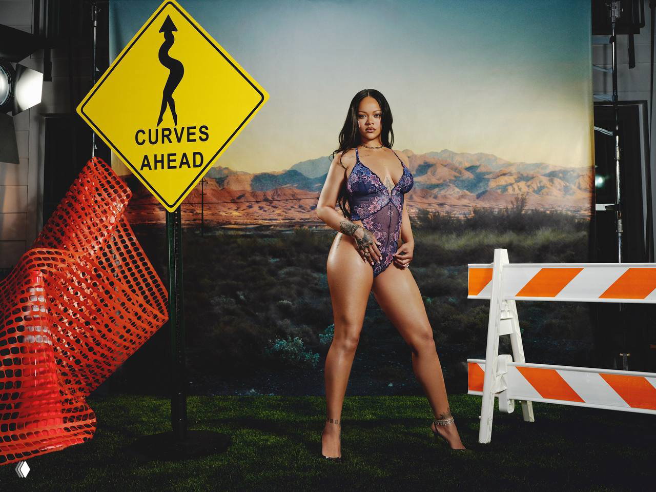 Широкий план съёмочной сцены: Рианна в боди Savage x Fenty на фоне дорожных барьеров и знака, студийный пустынный пейзаж.