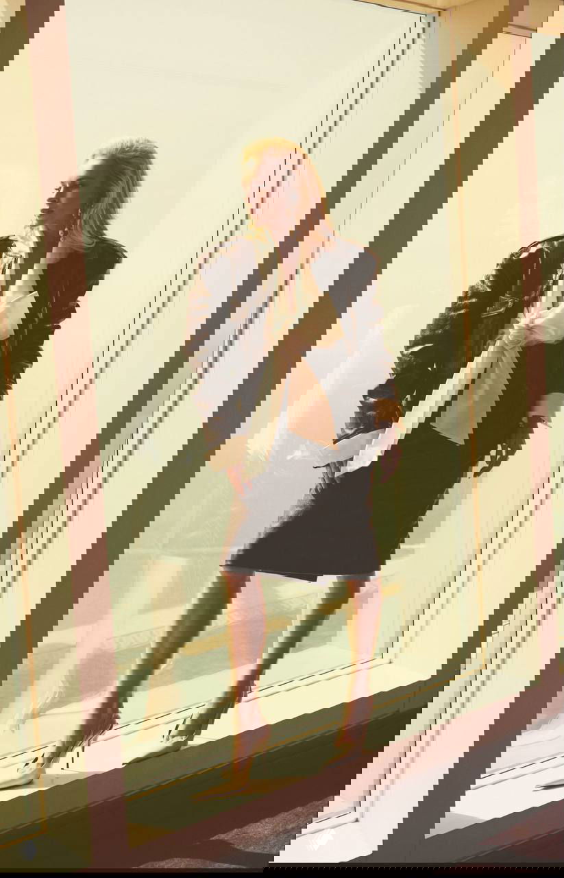 Линдси Лохан в кожаной куртке и блузе с бантом стоит у панорамного окна, выдержанное светлое модное фото с отражением в стекле.