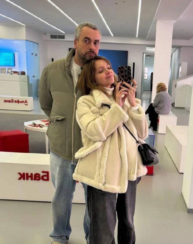 Селфи в галерейном интерьере: женщина в светлой дублёнке делает фото на телефон, рядом высокий мужчина, выставочные объекты и минималистичный интерьер.