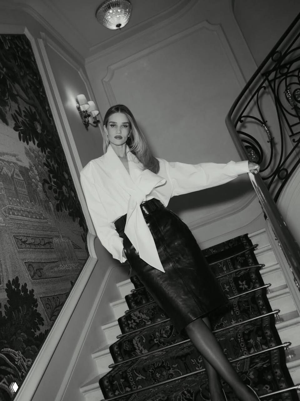 Чёрно-белый кадр: Роузи Хантингтон-Уайтли на лестнице в белой блузе и кожаной юбке, ретро-атмосфера и драматичный ракурс.
