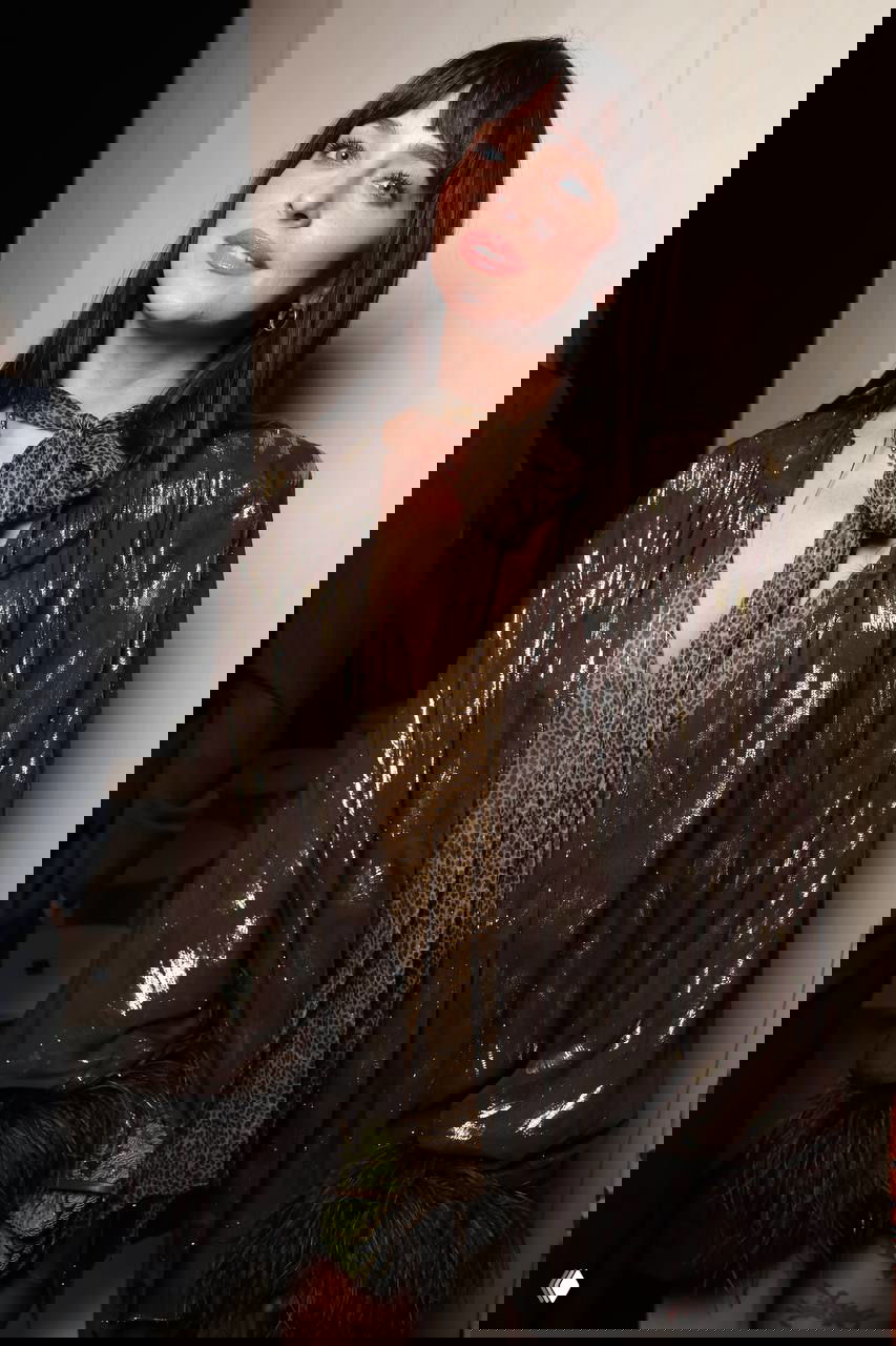 Крупный план лица женщины с тёмной челкой в мерцающем жакете и леопардовым шарфом на светском мероприятии.