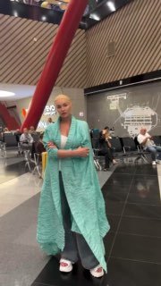 Та же женщина в бирюзовом наряде стоит в зале ожидания аэропорта на фоне кресел и архитектурных элементов терминала; рядом другие пассажиры.