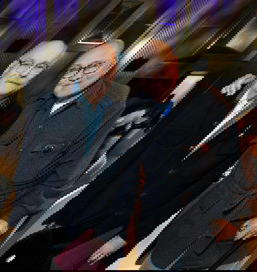 Два пожилых мужчины стоят рядом, улыбаются в камеру — кадр дружеской встречи коллег и близких на юбилее.