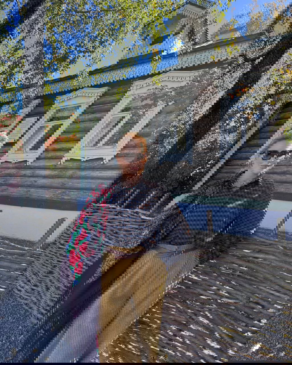 Женщина в тёмной клетчатой рубашке и вельветовых брюках стоит у плетёной изгороди на фоне старинного деревянного дома и берёз.