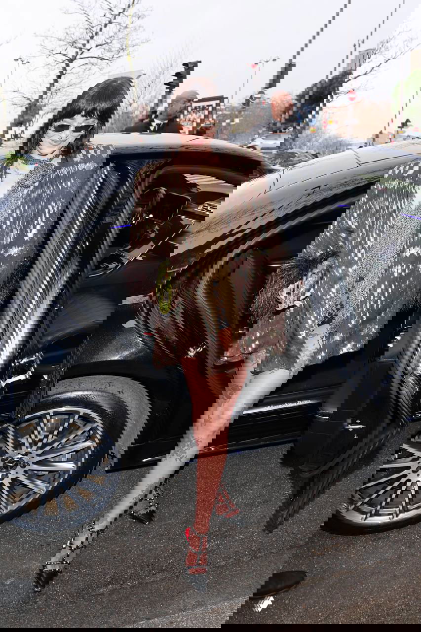 Женщина выходит из автомобиля в мини-образе и чулках с узором, на фоне городской улицы и охраны показа мод.