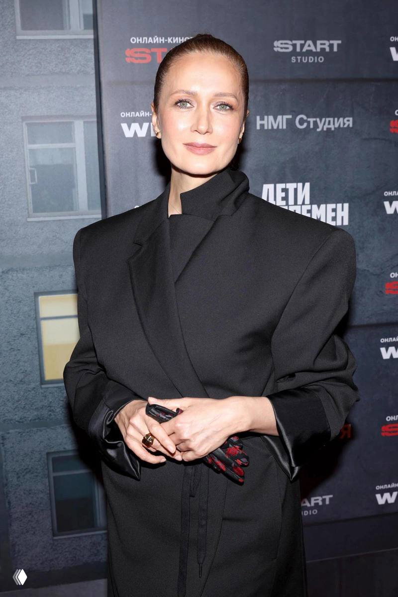 Женщина в чёрном пальто с аккуратной причёской позирует на фоне брендированного баннера кинопремьеры, официальный вечерний образ.