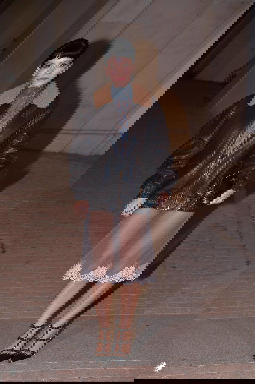 Героиня в коричневой тяжёлой кожаной куртке с меховым воротником и прозрачной юбкой с кружевной отделкой позирует на дорожке мероприятия.