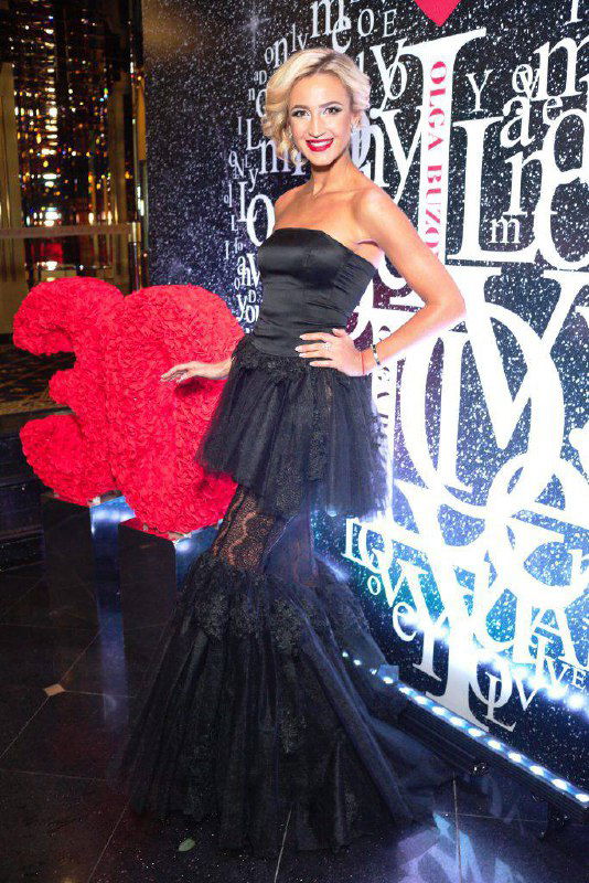 Женщина в чёрном вечернем платье с пышной многослойной юбкой и открытыми плечами на фоне фотозоны, вариант наряда для гала-вечера.