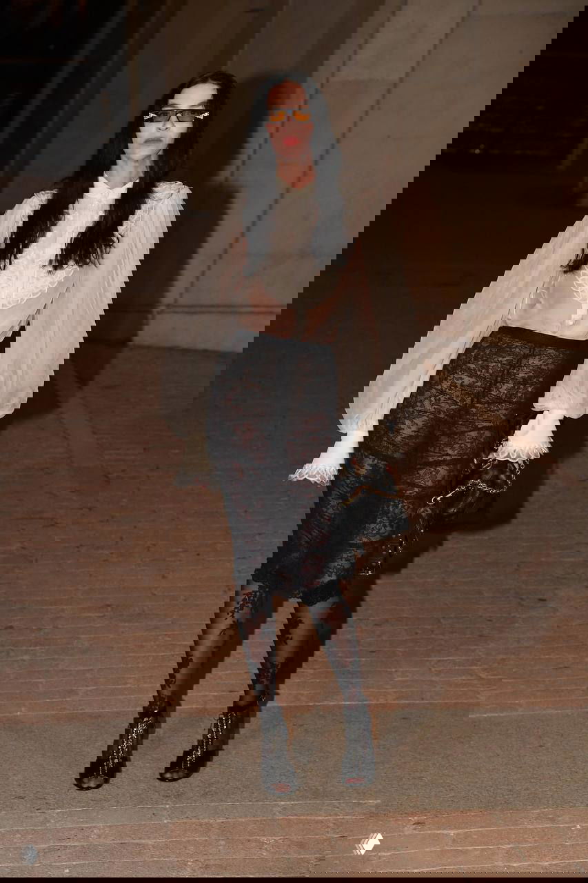 Женщина в белой прозрачной блузе с объёмными рукавами и чёрной кружевной юбкой идёт по красной дорожке показа мод вечером.