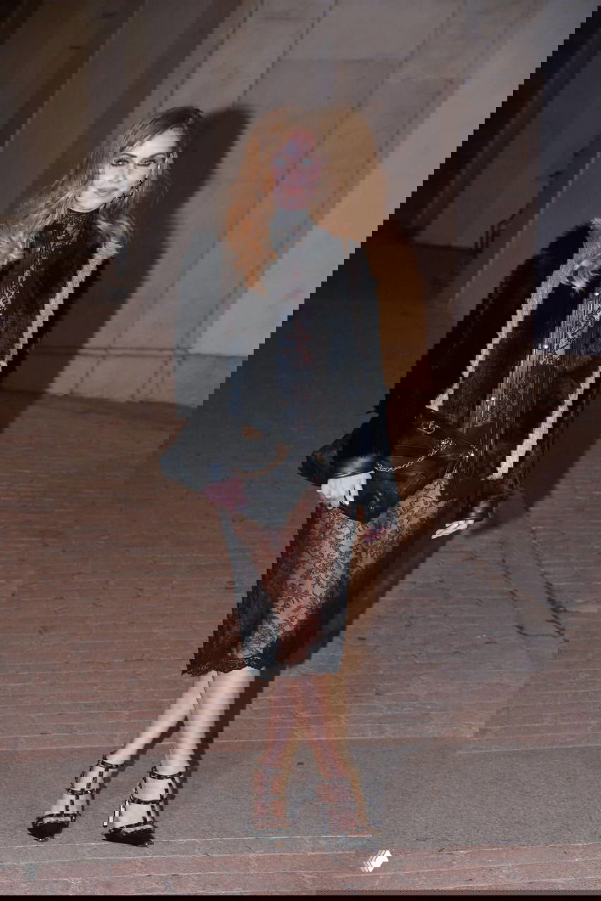 Портрет женщины в тёмном длинном платье с кружевной отделкой и чёрной сумкой, снятый на фоне колонн и кирпичной дорожки в сумерках.