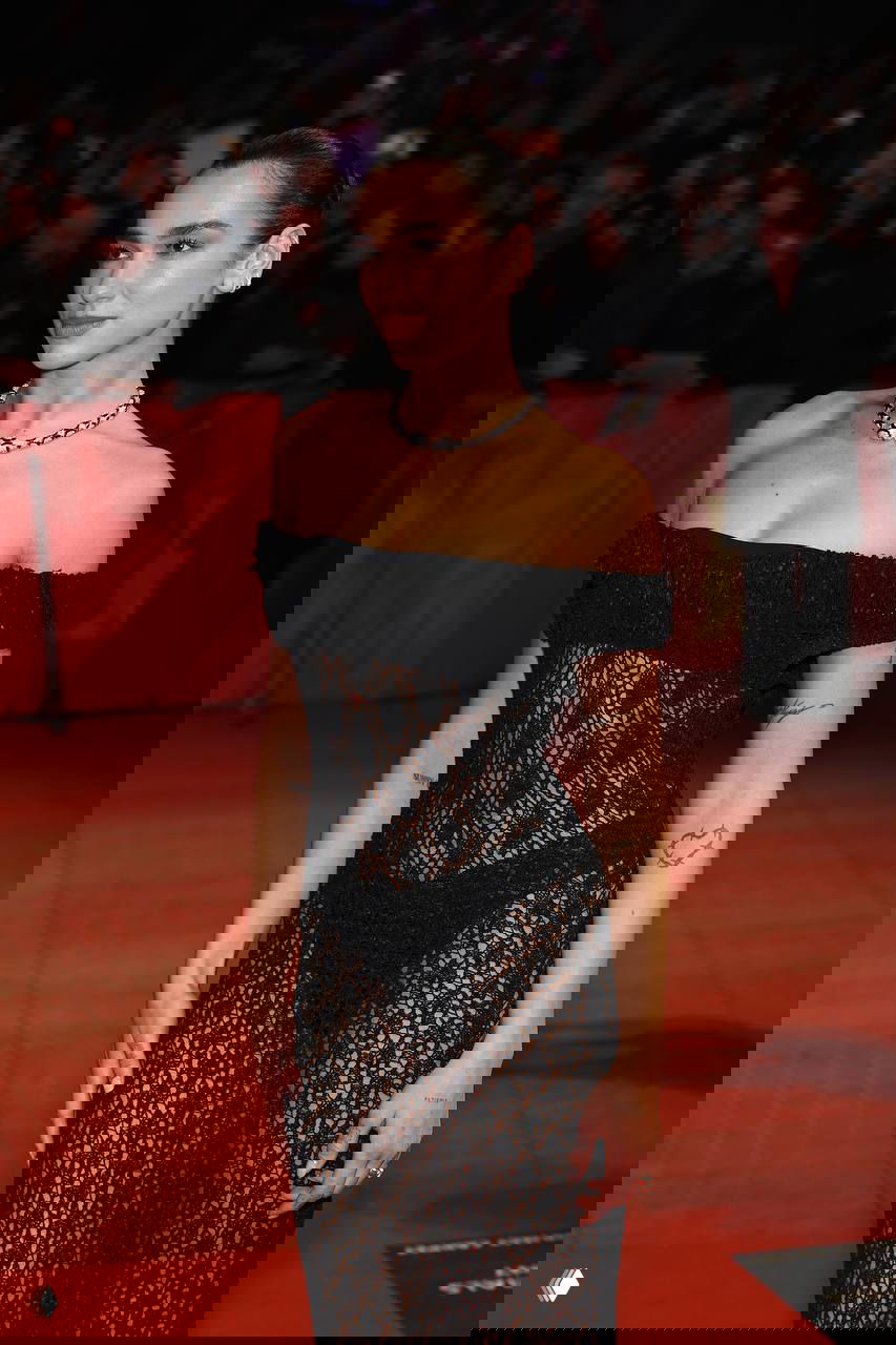 Крупный план певицы в чёрном прозрачном платье на красной дорожке, видны украшения и макияж, за ней толпа фотографов.