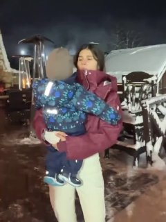 Кадр на открытой тёплой веранде: женщина держит ребёнка в тёплой одежде, вокруг заметна зимняя ночь и украшенная территория с огнями.