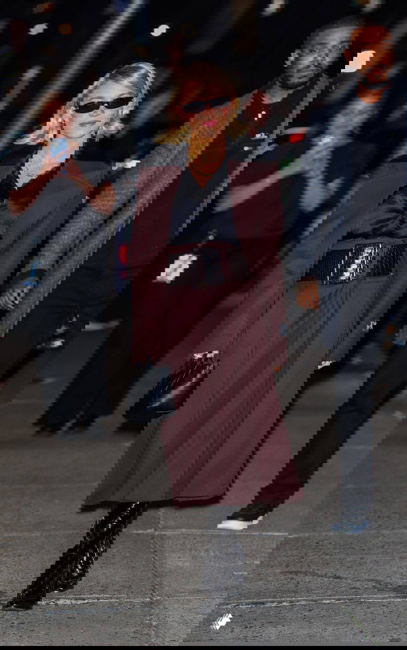 Фигуристая женщина в бордовом пальто и широком поясе, чёрная блуза и высокие лакированные ботильоны на городском тротуаре вечером.