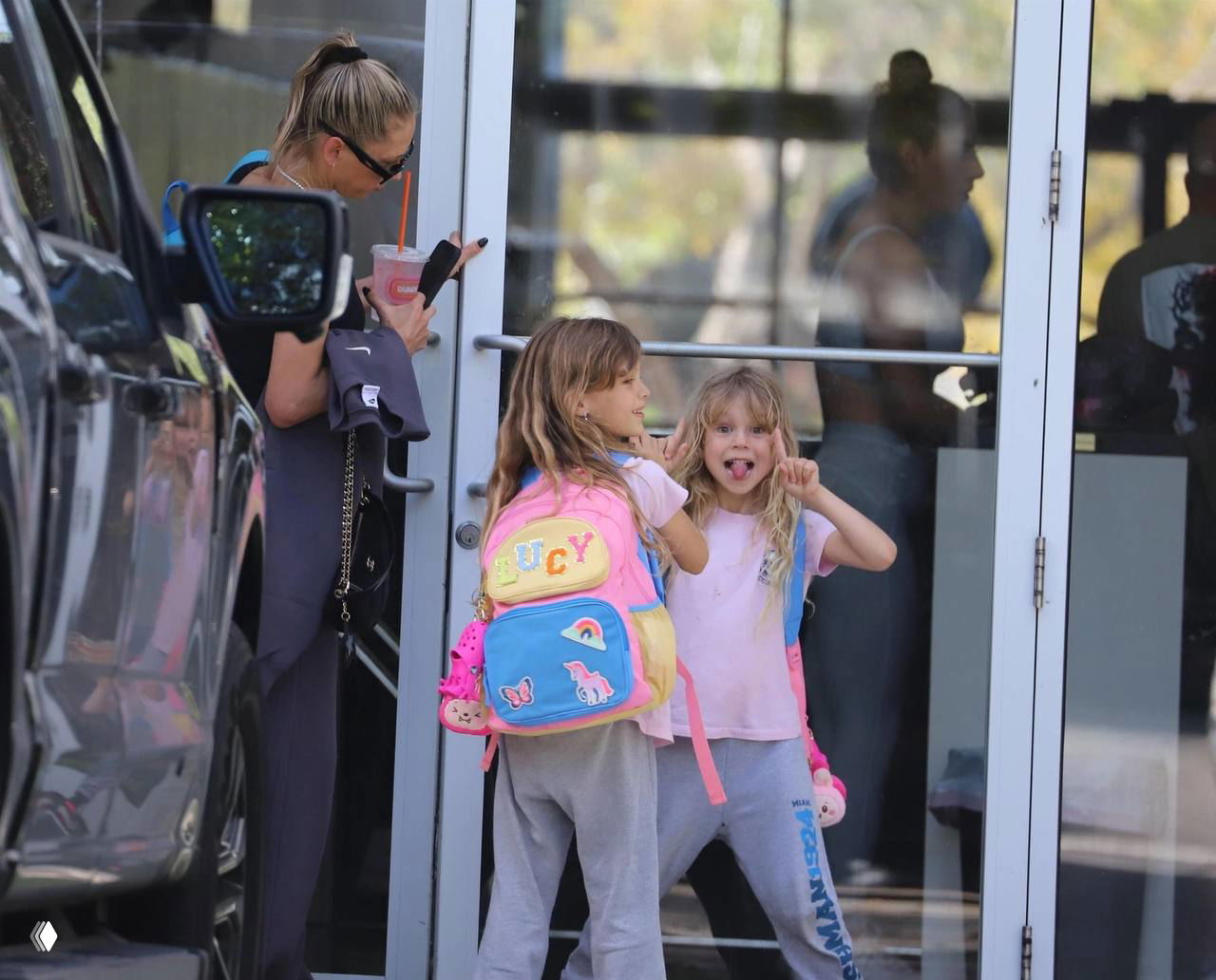 Две девочки в розовых футболках и рюкзаках стоят у двери здания; младшая демонстрирует язык и строит забавные рожицы, старшая рядом.