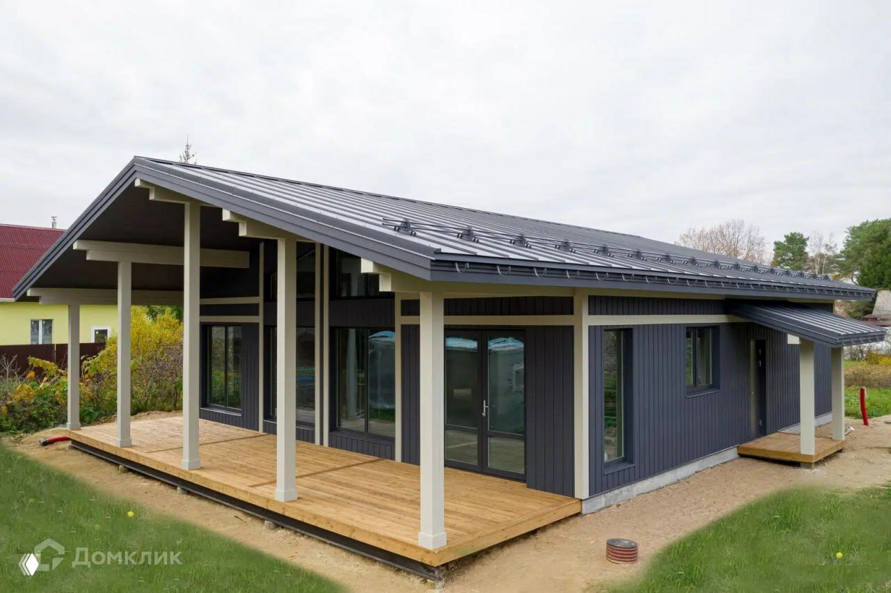 Современный одноэтажный дом с широкой террасой и тёмной кровлей: деревянный настил, большие панорамные окна и газон вокруг.