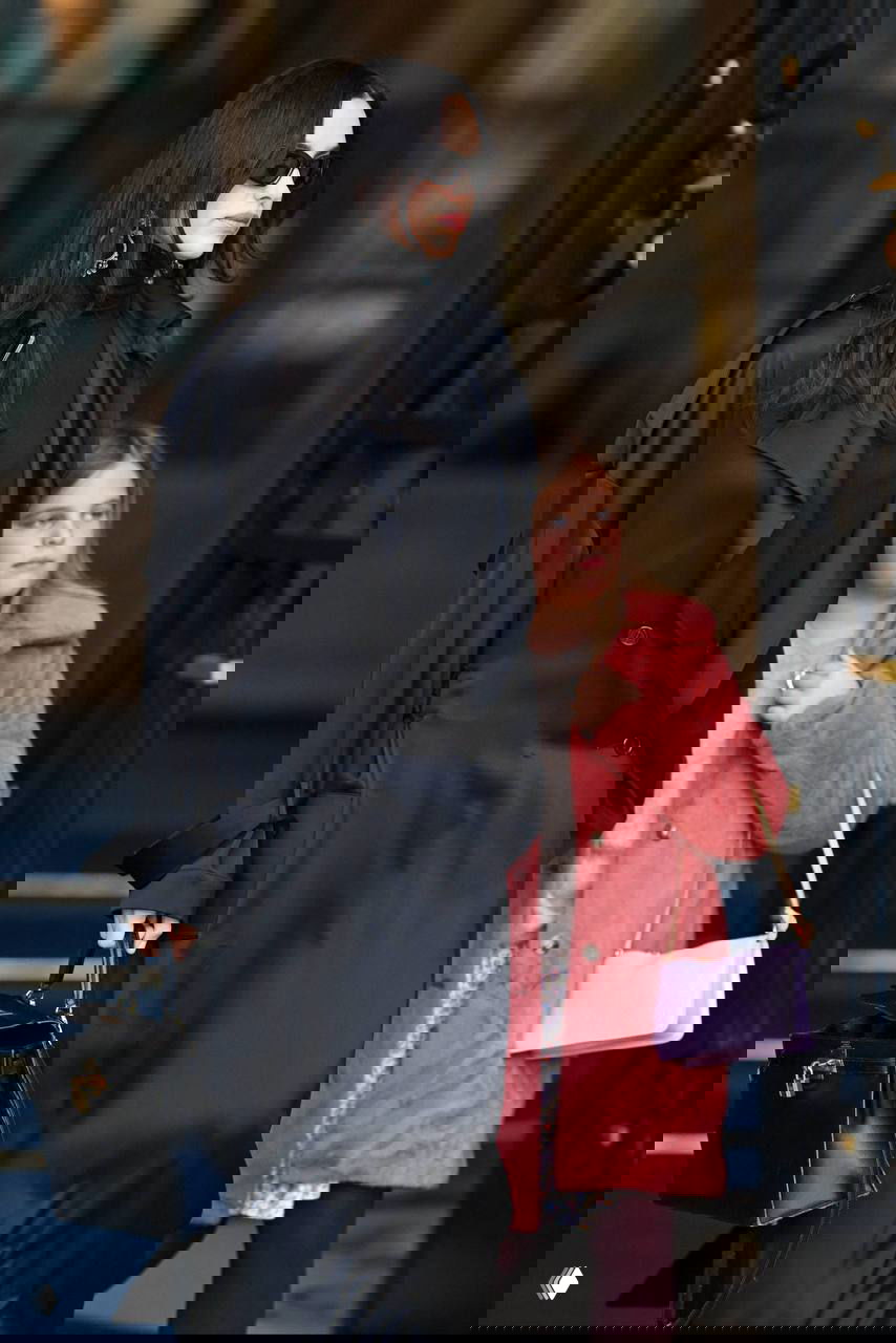 Женщина в эффектном чёрном образе идёт рядом с девочкой в розовом пальто у входа в здание в Париже; уличный папарацци-кадр.