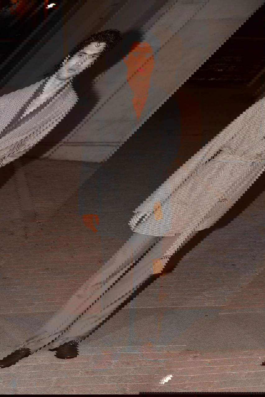 Мужчина в сером костюме с поясом и свободными брюками позирует на открытой площадке возле архитектурных колонн на вечернем шоу моды.