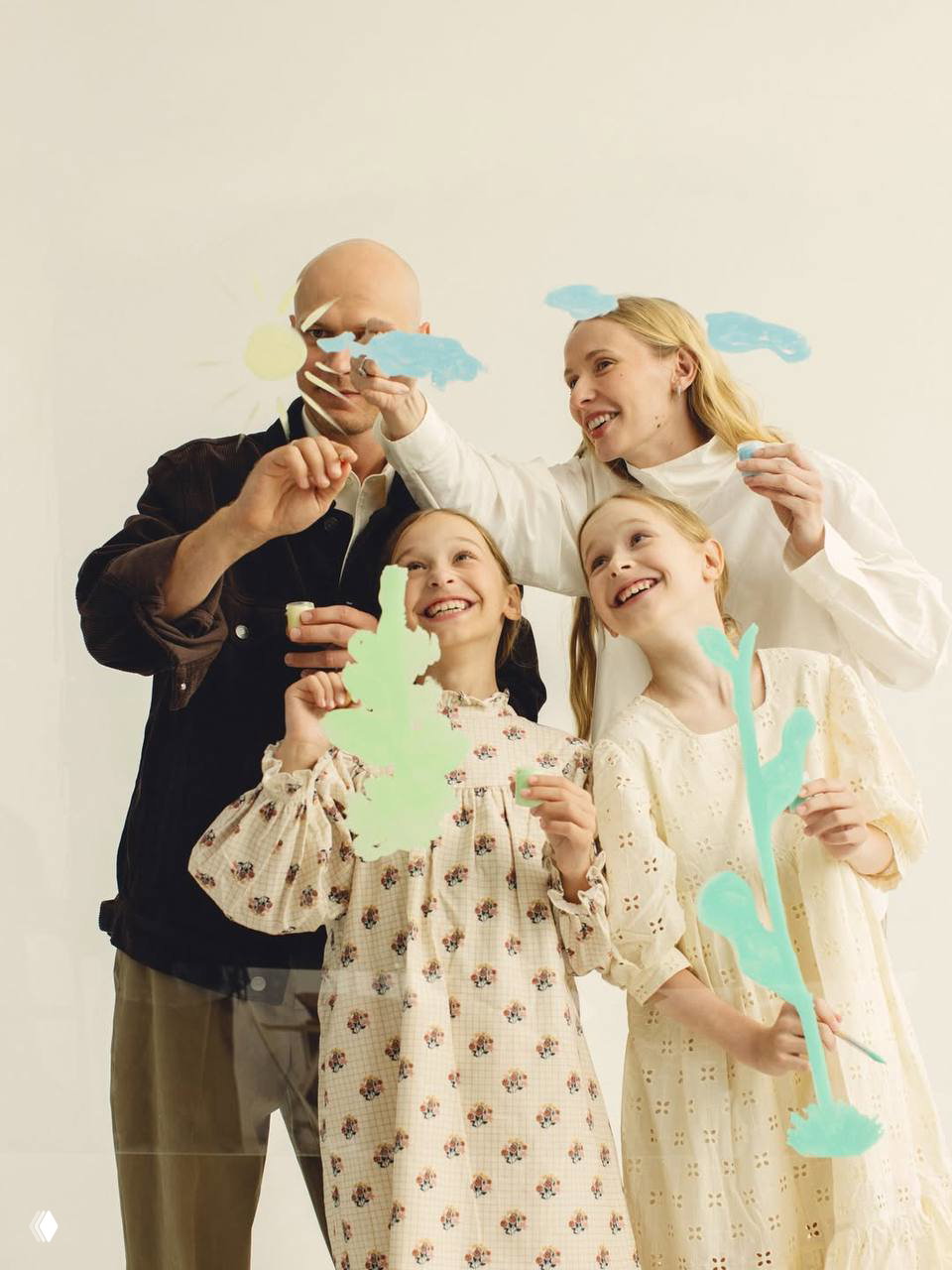 Семья улыбается и держит бумажные декорации в руках: тёплая студийная сцена с родителями и двумя дочерьми в пастельных тонах.