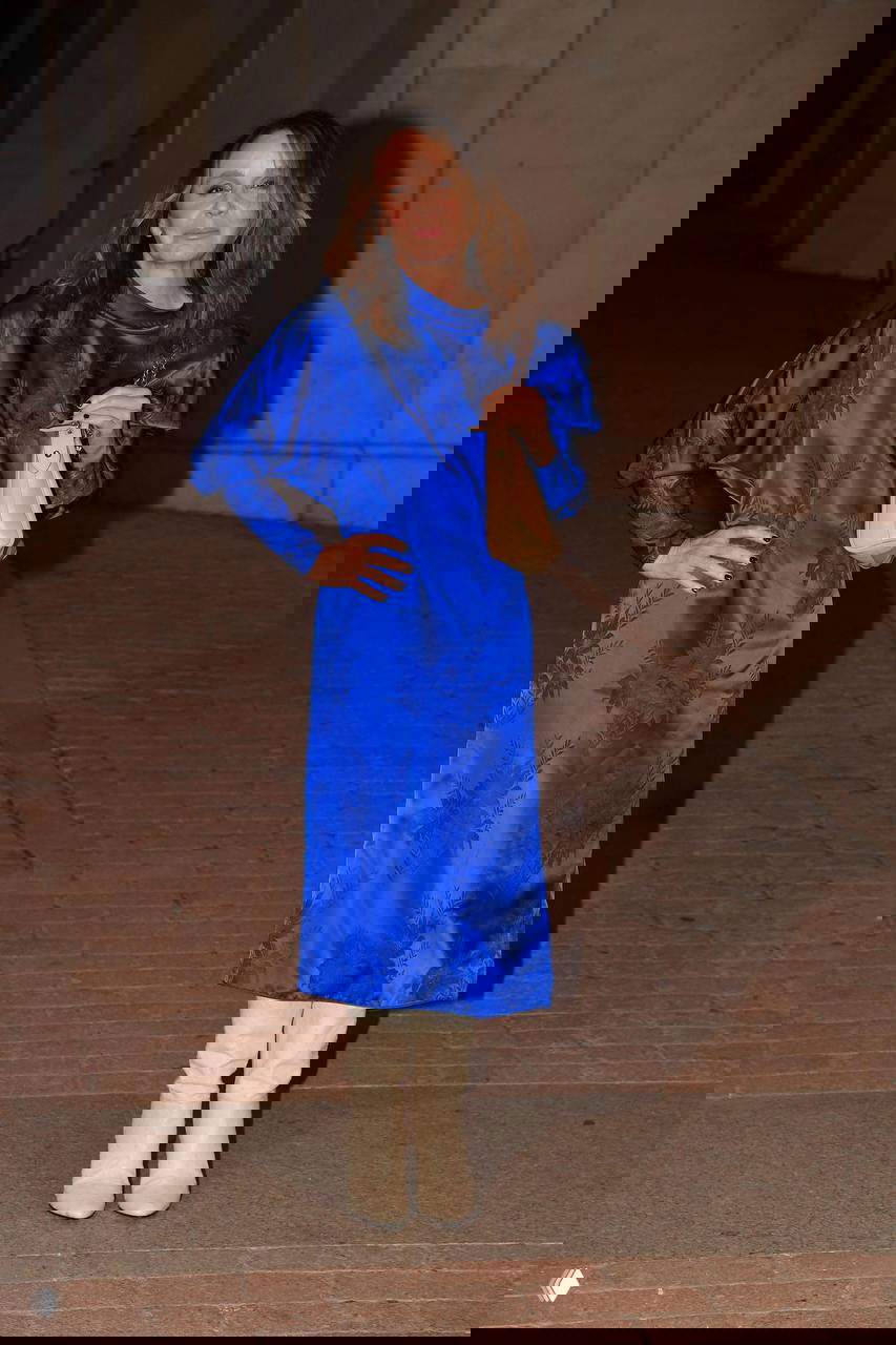 Женщина в длинном ярко-синем платье с узорчатой текстурой и светлыми ботинками позирует на кирпичной дорожке у фасада здания.