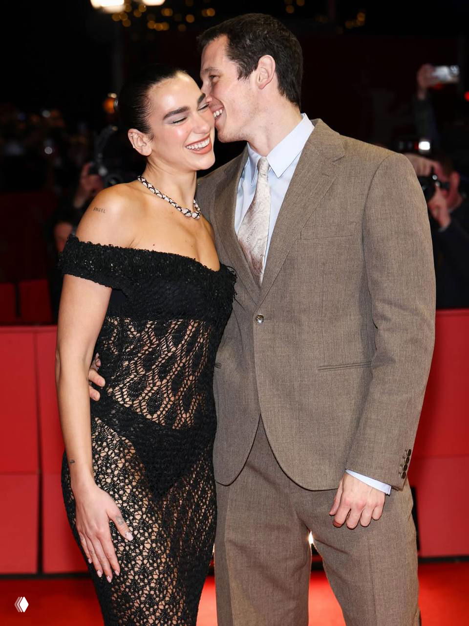 Певица и мужчина в костюме обнимаются на красной дорожке; платье остаётся прозрачным, вокруг фотографы и фирменные баннеры фестиваля.