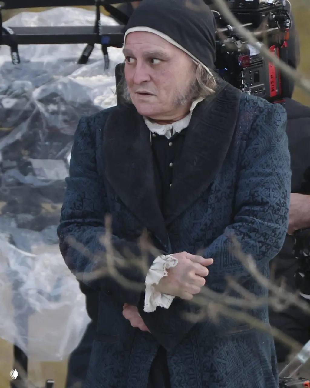 Крупный план актёра в историческом костюме и чёрной шапочке на фоне киносъёмочной техники; кадр сделан на открытой площадке, видны детали грима.