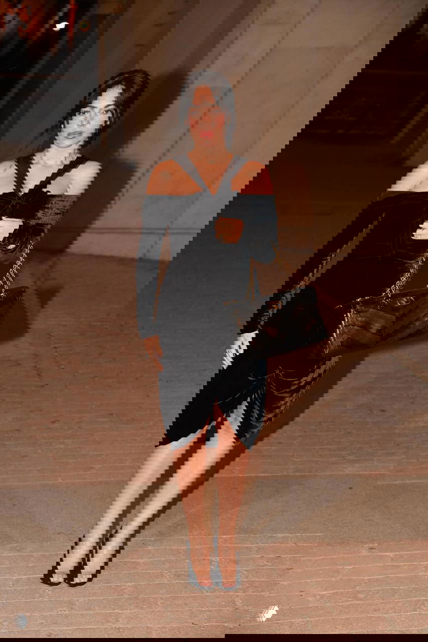 Женщина в чёрном бархатном платье с открытыми плечами и большой сумкой проходит по дорожке под вечерним освещением на показе.