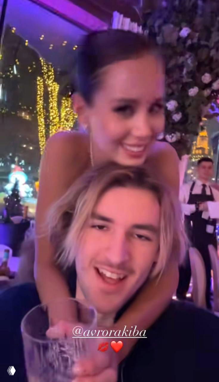 Крупный план гостей: молодая женщина сидит на плечах мужчины, на заднем плане праздничная иллюминация и официанты, атмосфера вечеринки.