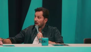 Мужчина в тёмной рубашке сидит за столом и говорит в микрофон; на столе кружка и бумаги, фон — зелёный.