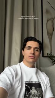 Луис в белой футболке делает видео‑сообщение в домашней обстановке: на заднем плане занавески, картина и комнатное растение, интерьер в нейтральных тонах.