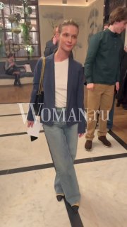 Женщина в пиджаке и джинсах в фойе кинотеатра на премьере документального сериала; люди и декор на заднем плане