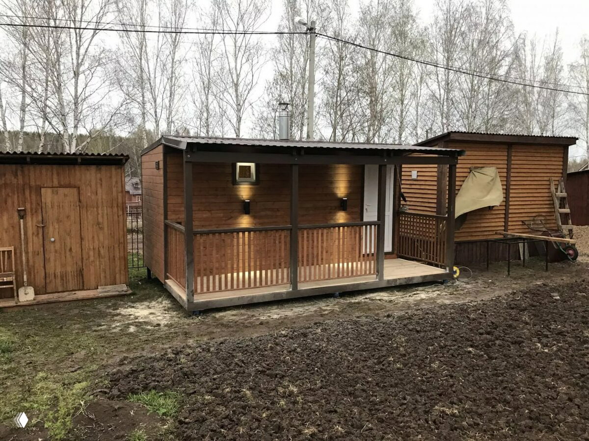 Небольшой деревянный дачный домик с крытой верандой и настенной подсветкой на фоне огороженного садового участка и голых весенних деревьев.