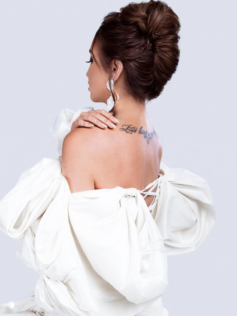 Кадр со спины: женщина в платье с открытой спиной, видна небольшая татуировка на лопатке и аккуратная причёска, студийный минималистичный фон.