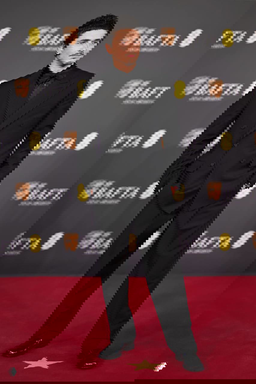 Тимоти Шаламе в чёрном смокинге с бабочкой на красной дорожке BAFTA‑2026 в окружении брендинга и фонового баннера.